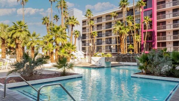 A hotel with a pool surrounded by palm trees, a multi-story building with balconies, and a clear blue sky. - South Country Suites (Mesa)