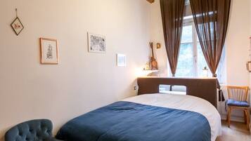 Een slaapkamer met een houten plafond, een bed met een blauwe spreidekking, een ronde stoel en een raam met gordijnen.