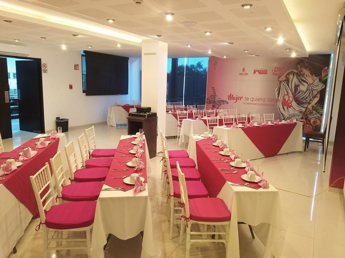 Une salle de banquet avec des rangées de tables dressées pour un événement formel, une scène avec un décor en fond et une grande fresque murale.
