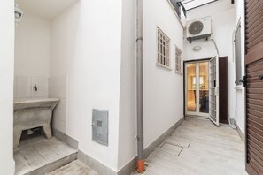 A narrow hallway with a bathroom on the left, a door with a view into a room on the right, and a window with an air conditioning unit above it.