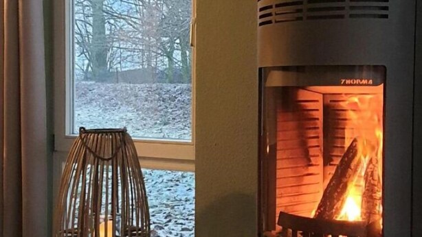 Een houtkachel met een zichtbare vlam, een raam met uitzicht op kale bomen en een geweven kandelaar op de vloer.