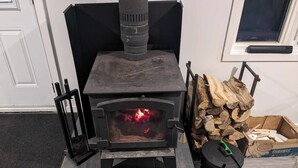 A wood-burning stove with a visible fire, a stack of firewood, and a black trash bin. - Chalet Rustique sur île (Saguenay)