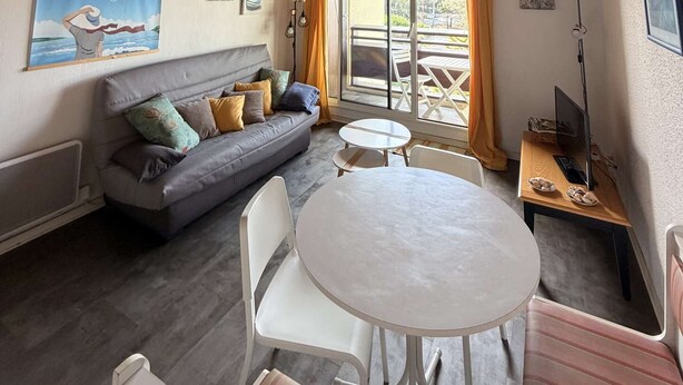 Et hyggeligt værelse med træloft, en grå sofa med puder, et rundt hvidt bord og udsigt til en udendørs terrasse.