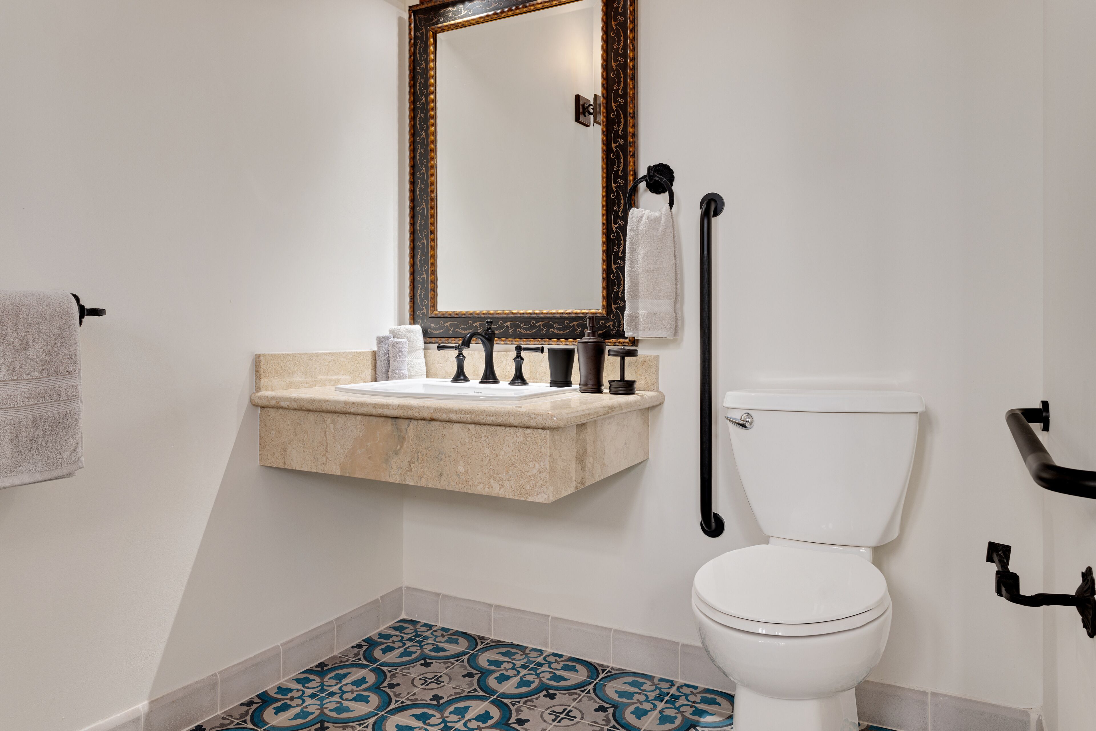 Une salle de bain avec des toilettes blanches, un lavabo beige avec un miroir au-dessus, et des carreaux de sol à motifs.