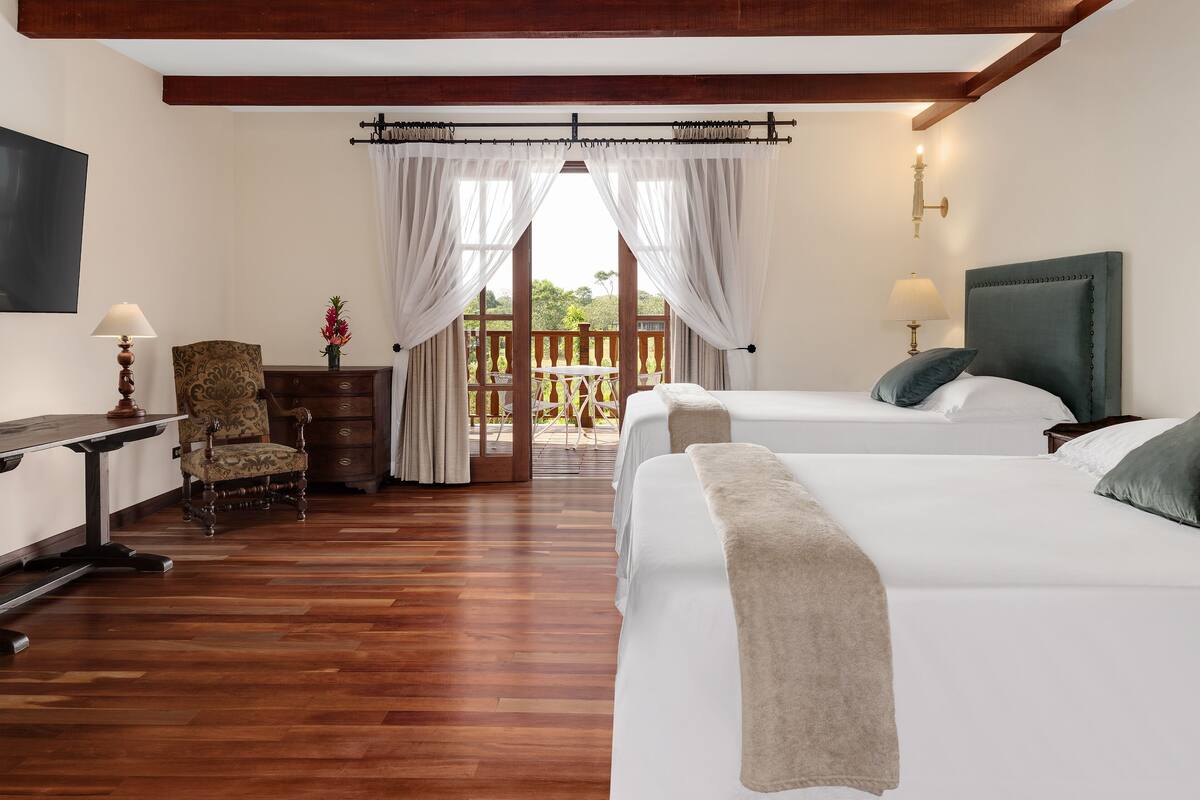 A hotel room with two beds, wooden flooring, a desk with a lamp, a chair, and a view of a balcony.