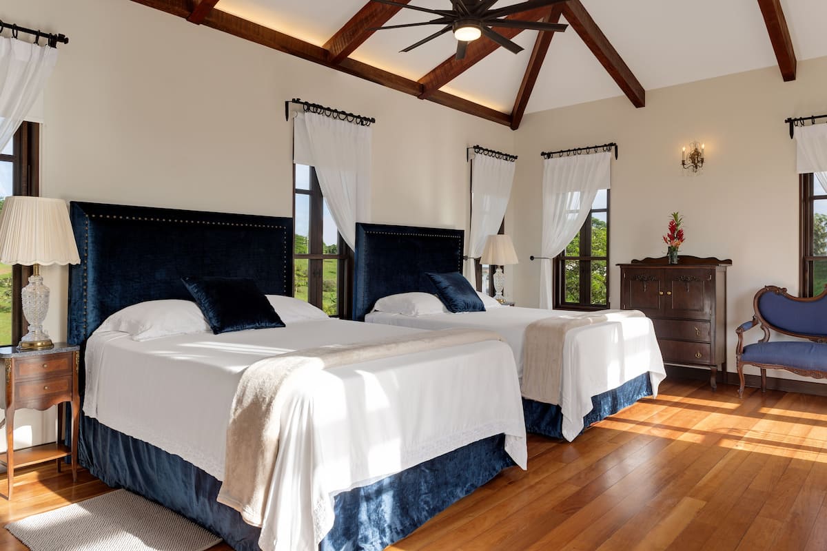 A bedroom with two beds, wooden flooring, a ceiling fan, and a nightstand with a lamp.