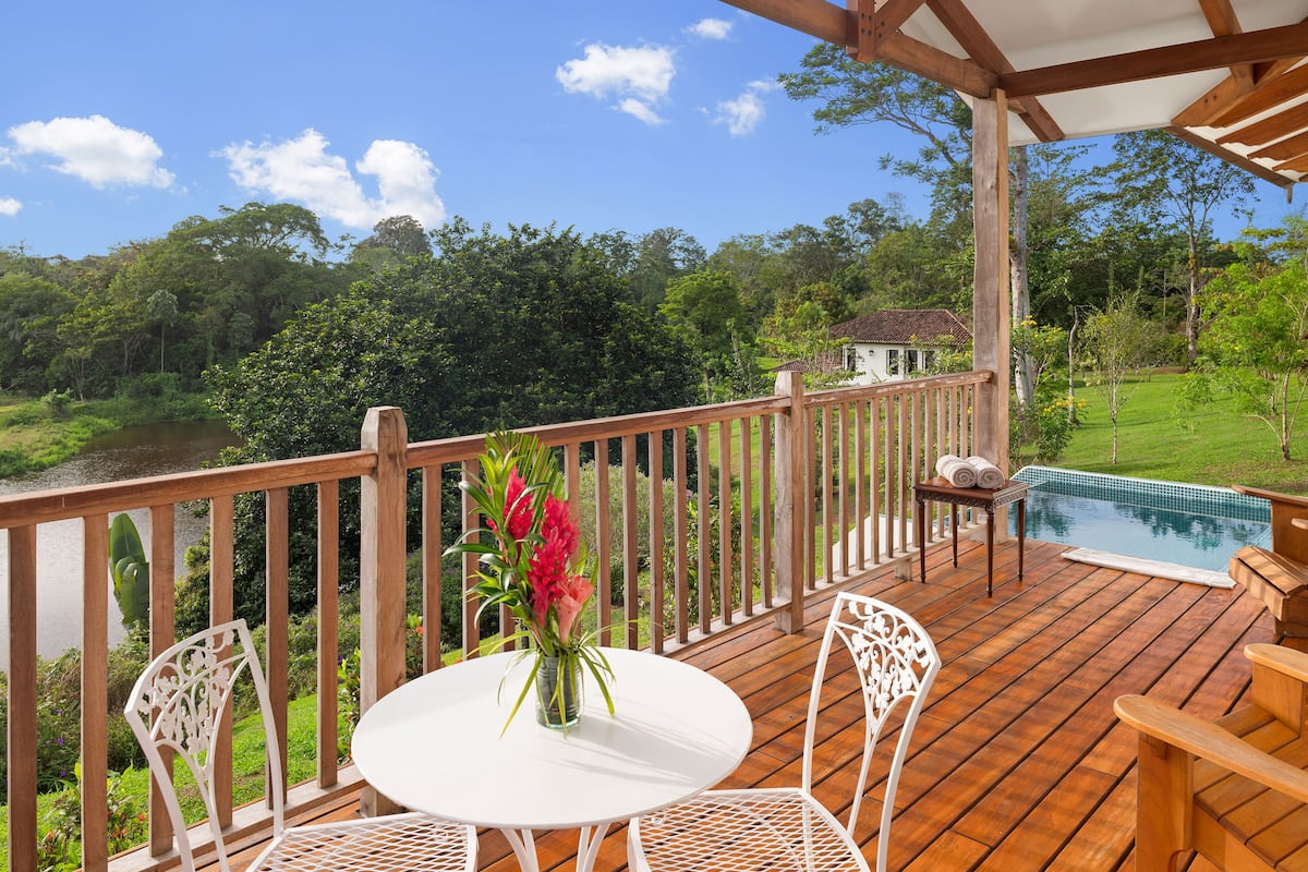 A wooden deck with a white table and chairs, overlooking a swimming pool and lush greenery.