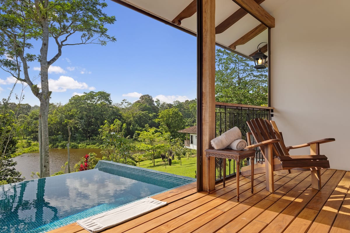 A wooden deck with a pool, a lounge chair, and a view of a lake and greenery.