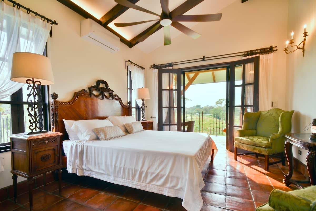 A bedroom with a wooden bed, a nightstand, a green armchair, and a view through a large window.