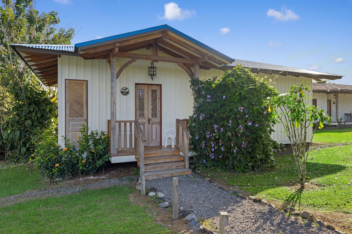 A white building with a wooden porch, a door, and a small garden with flowers and a tree.