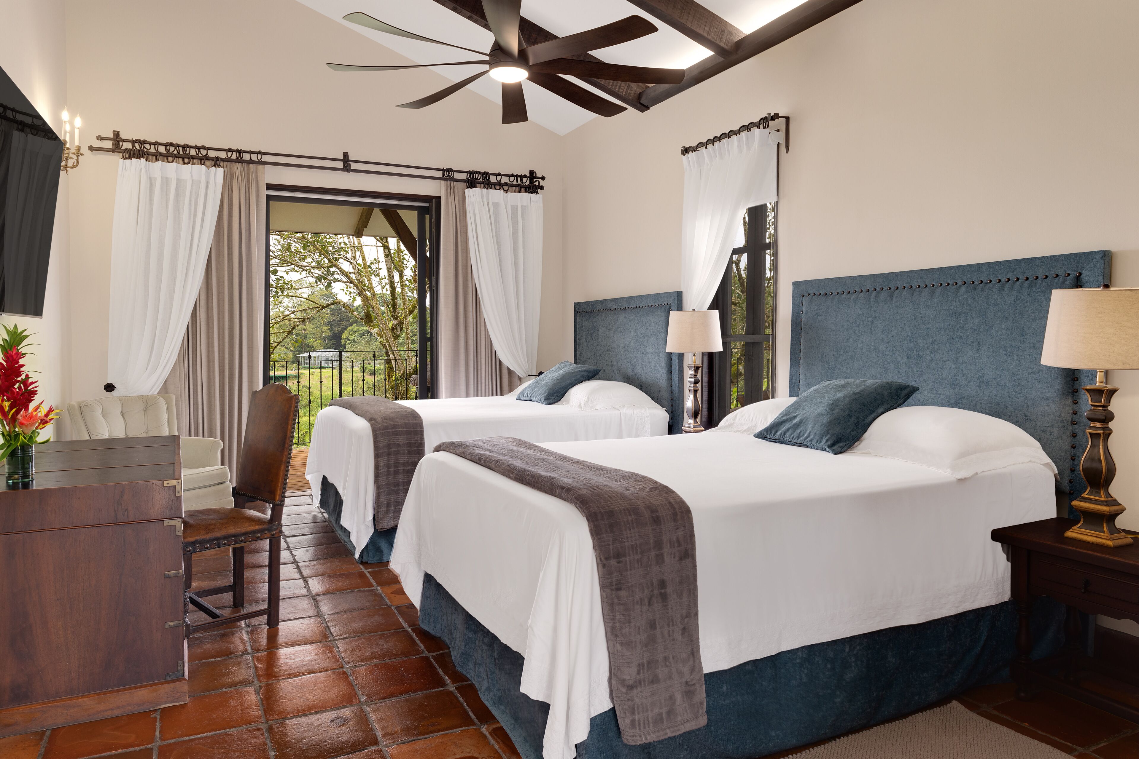 A hotel room with two beds, a desk, a chair, and a ceiling fan.