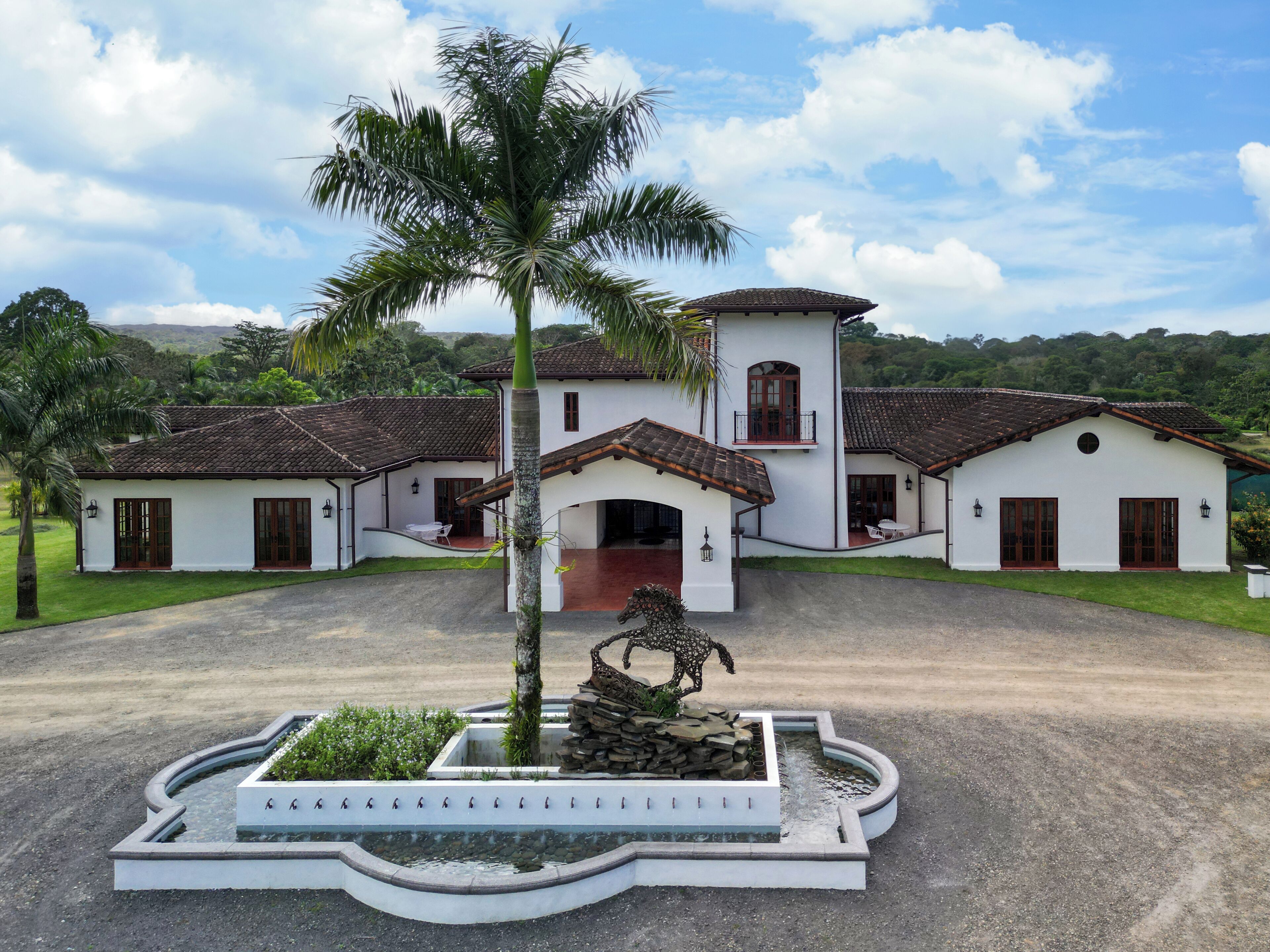 A large, white building with a tiled roof, surrounded by lush greenery and a clear sky.