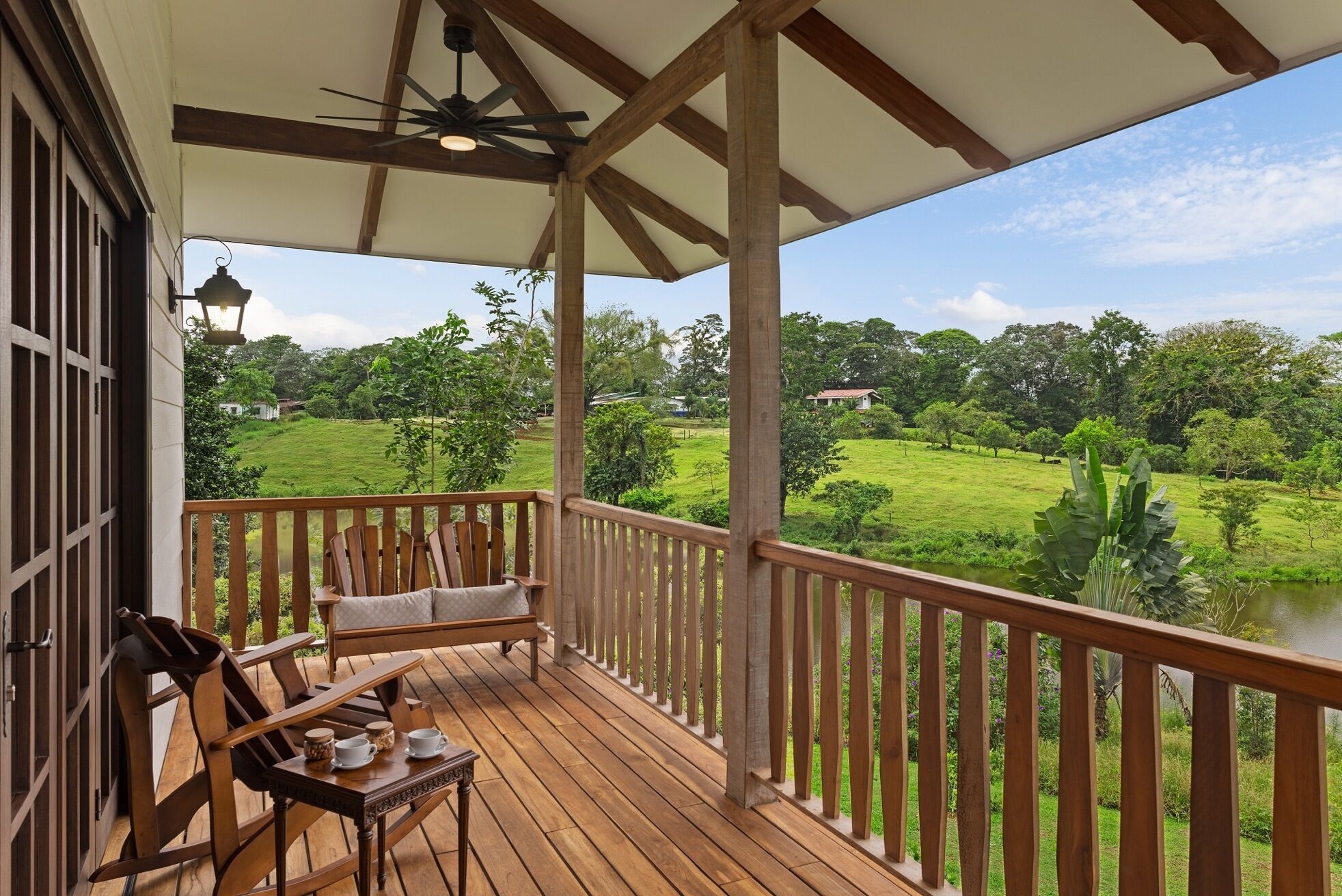 A wooden deck with a chair and table, a ceiling fan, and a view of a green landscape with trees and a house.