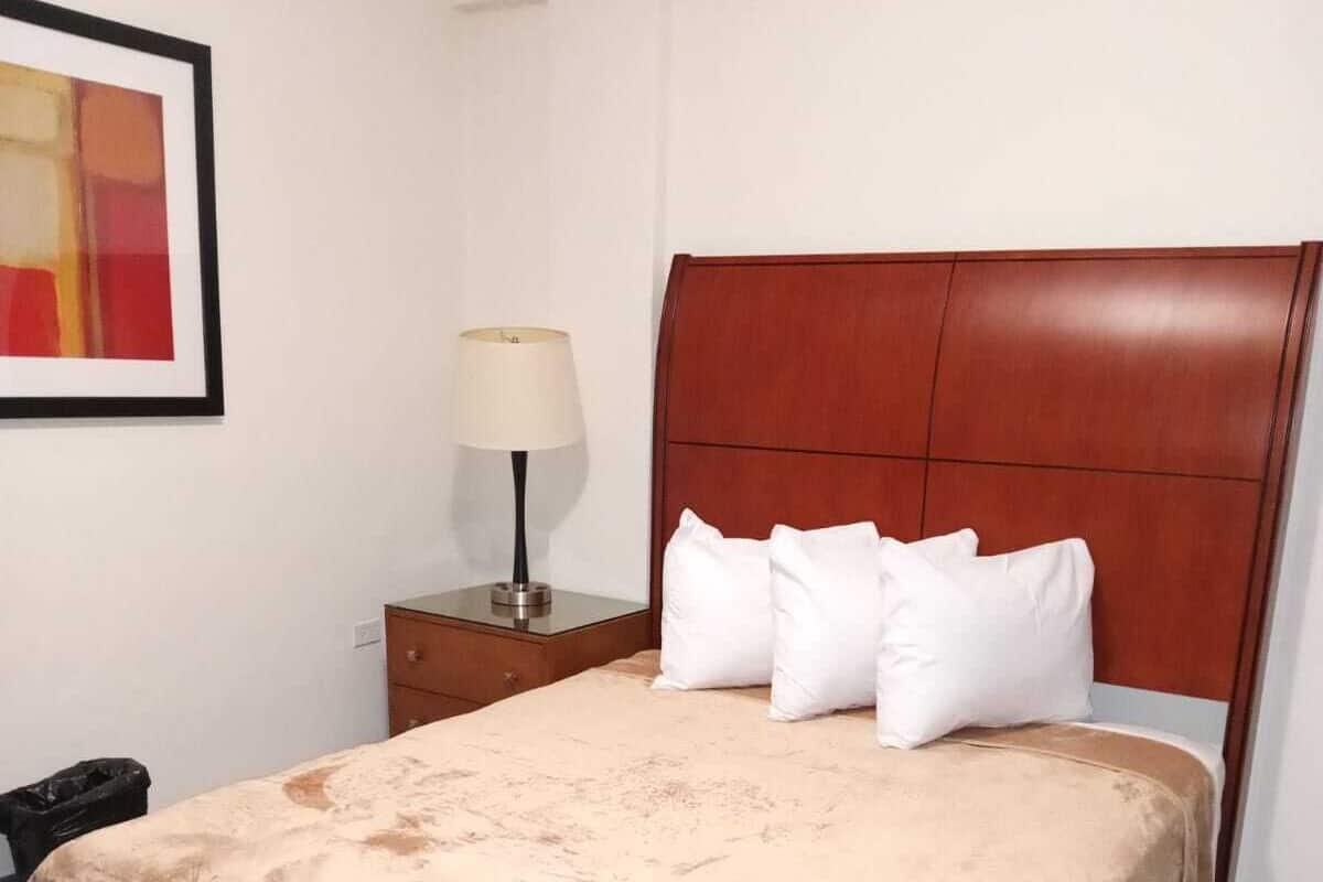 Una cama bien hecha con una colcha marrón, cabecera de madera y almohadas blancas. Hay una mesita de noche con una lámpara y un cuadro en la pared.