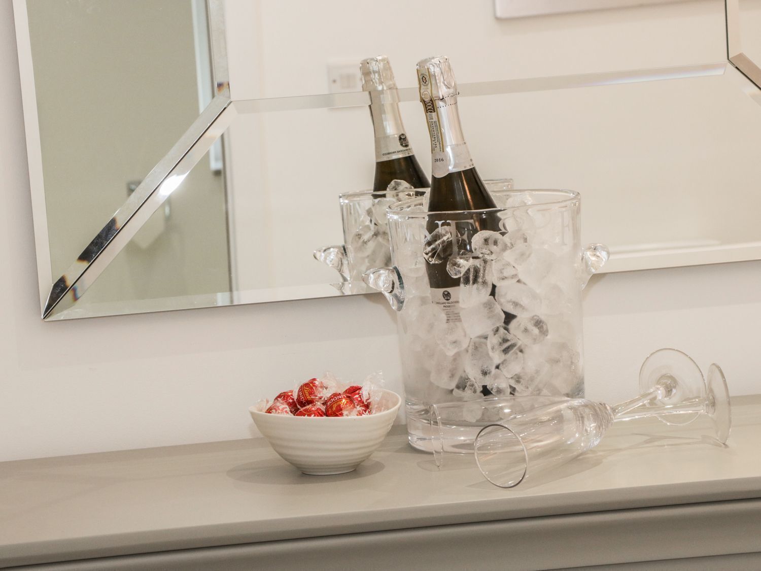 A clear glass ice bucket with two champagne bottles and a bowl of chocolates on a white countertop.