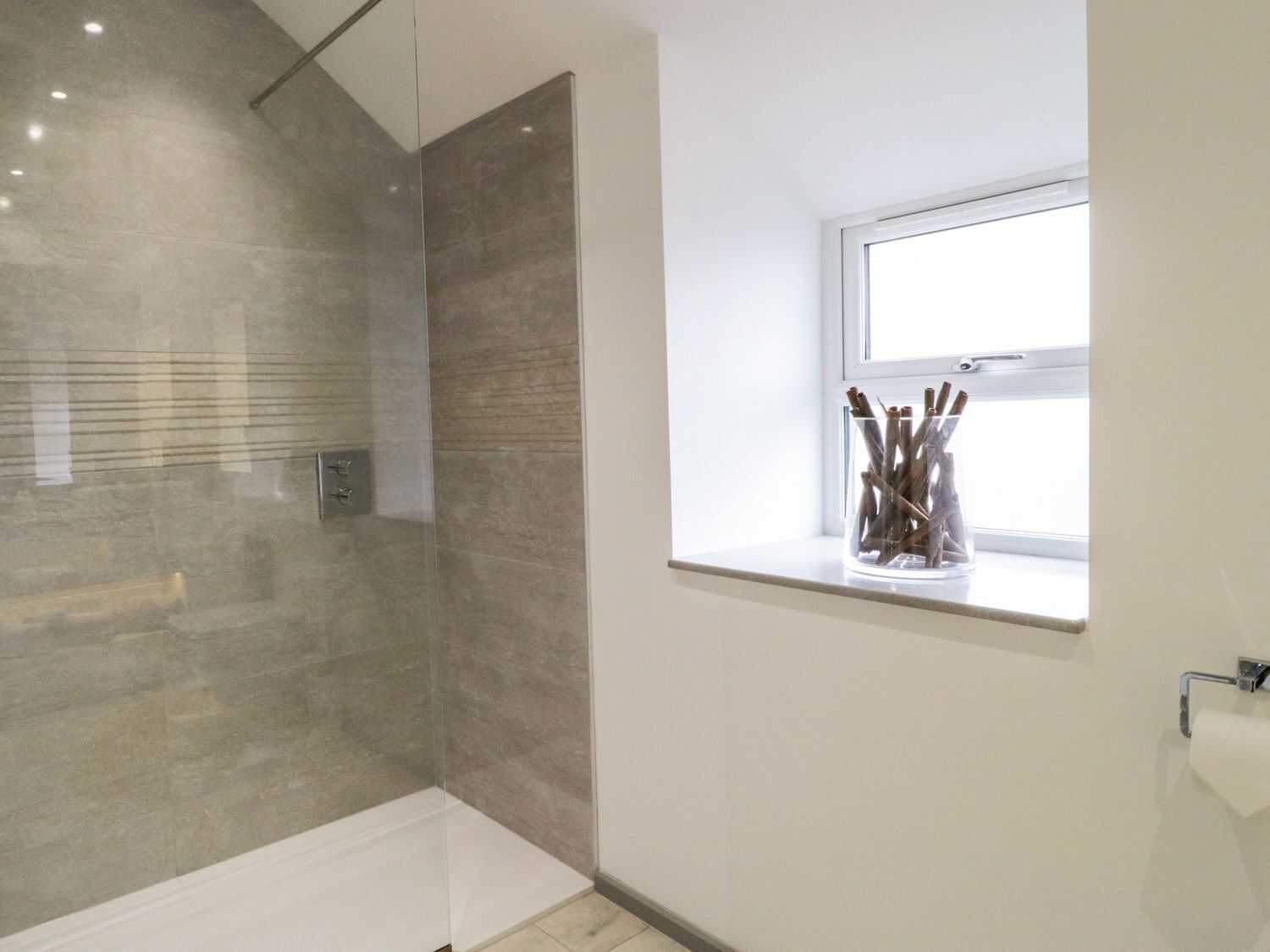 A modern bathroom with a glass shower enclosure, a window with a view, and a vase with sticks on the windowsill.