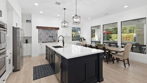 A modern kitchen with white cabinets, a large island, and a dining area with a view of greenery. - Serenity Retreat at Montage, by GPM - Pool + Gameroom + More! Walk to Festivals (Indio)