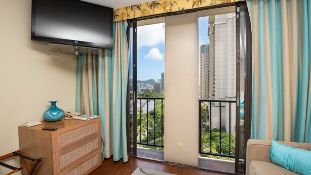 A living room with a flat-screen TV, a wooden dresser, a sofa, and a view of a cityscape through a balcony with striped curtains. - *Free Parking*Beautiful Views*10 Min to Beach* (Honolulu)