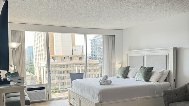 A modern hotel room with a large bed, a desk, and a view of the city through large windows.