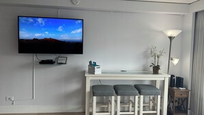 A modern hotel room with a flat-screen TV mounted on the wall, a small desk with a chair, a potted orchid, and a floor lamp.