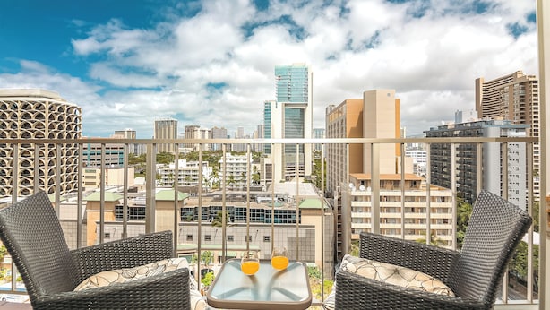 A balcony with wicker chairs and a table, overlooking a cityscape with various buildings and a clear sky. - Newly Updated*Free Parking*Full Kitchen*Close to Beach (Honolulu)