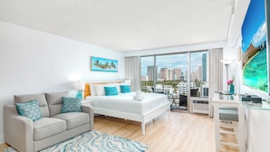 A modern hotel room with a bed, sofa, desk, and large windows offering a city view. - Newly Updated*Free Parking*Full Kitchen*Close to Beach (Honolulu)