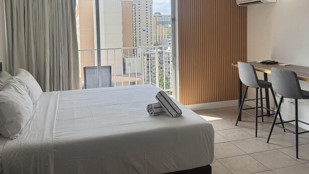 A hotel room with a bed, a desk, two chairs, a window with curtains, and an air conditioning unit. - King Size Bed*Free Parking*Walk to the beach (Honolulu)
