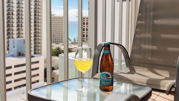 Een glas gele drank en een fles Big Wave bier op een balkon met uitzicht op hoge gebouwen en de oceaan.