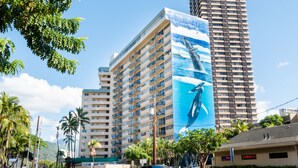 Un hotel con varios pisos, un llamativo mural con tema oceánico y vista a la calle con coches y palmeras.