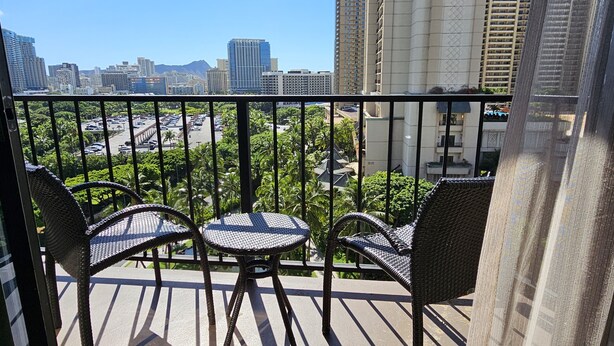 A balcony with wicker chairs and a table, overlooking a cityscape with tall buildings and a mountain in the distance. - Recently Renovated Studio in Palms at Waikiki (Honolulu)