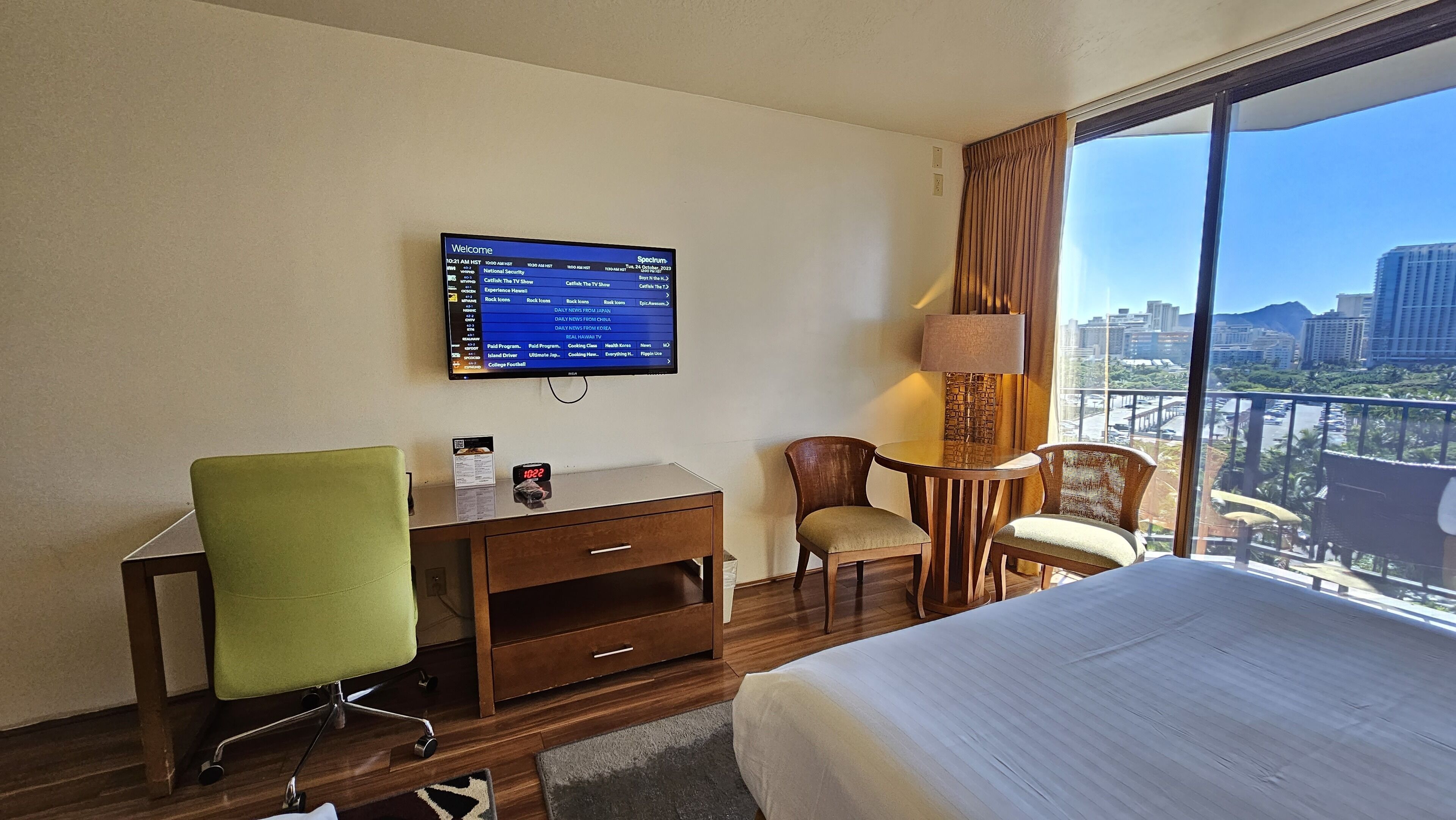 A hotel room with a bed, a desk, a chair, a TV displaying a program list, and a view of the city through large windows.