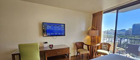 A hotel room with a bed, a desk, a chair, a TV displaying a program list, and a view of the city through large windows.