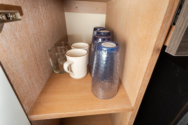 A wooden shelf with clear glasses, white mugs, and a blue glass container.