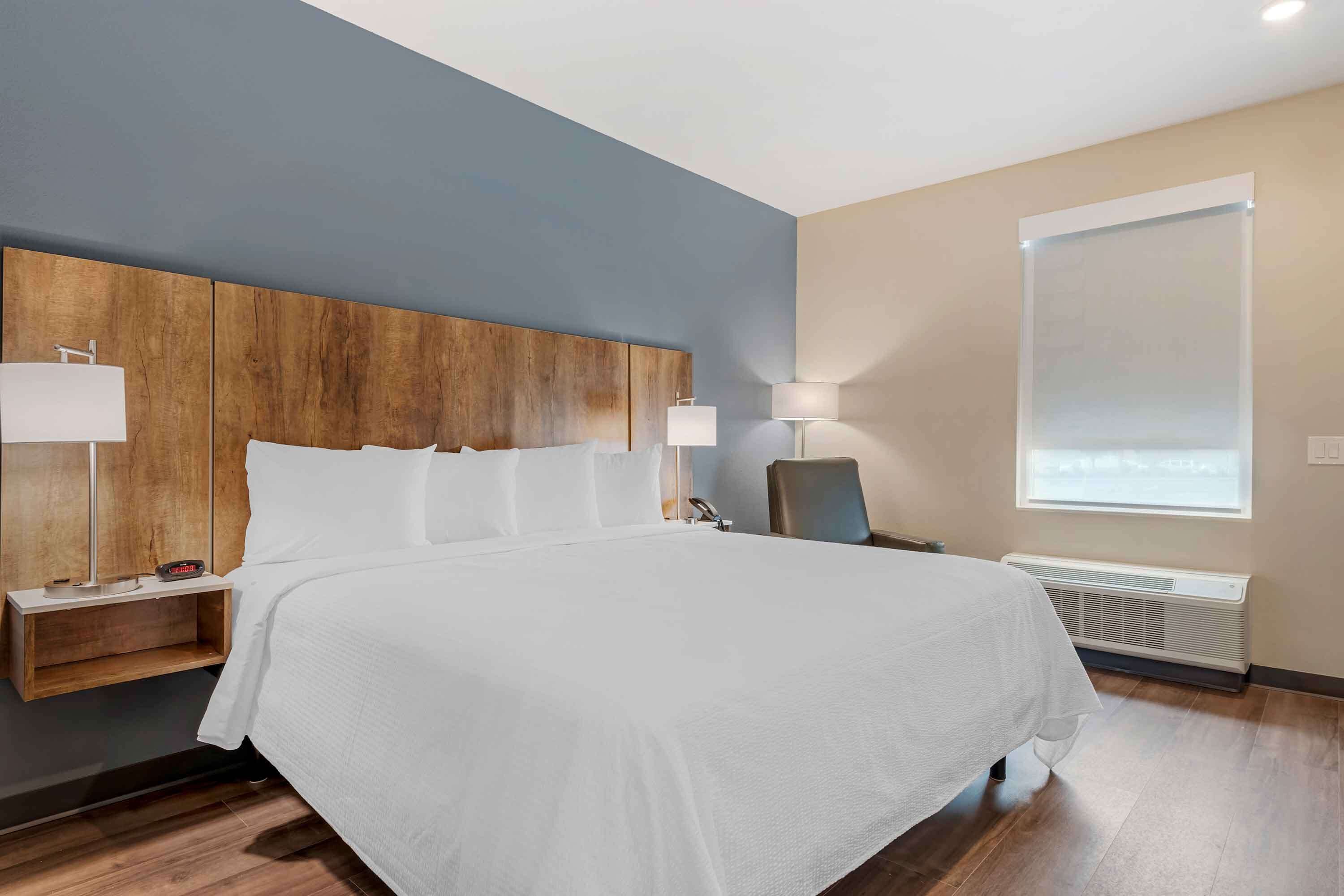A modern hotel room with a large bed, wooden headboard, bedside lamps, a chair, and a large window.