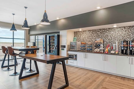 A modern kitchen with a wooden table, bar stools, pendant lights, a coffee station, and a view of the beach.