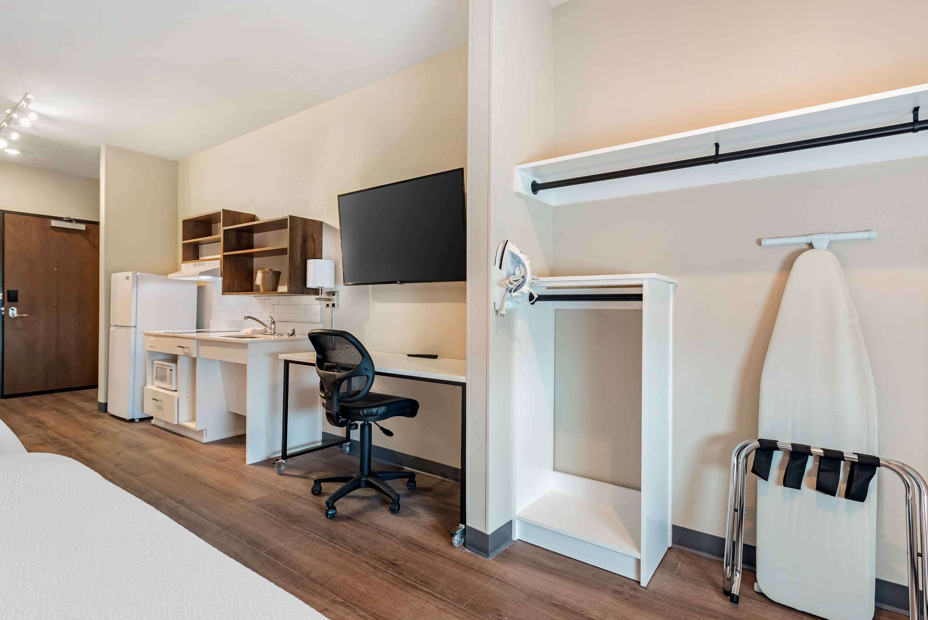 A modern hotel room with a wooden floor, a white bed, a desk with a chair, a flat-screen TV, a built-in ironing station, and a wardrobe.