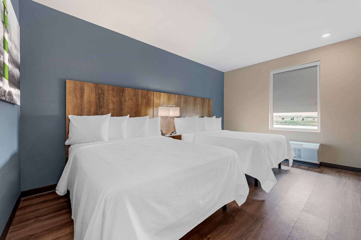 A hotel room with two beds, a wooden headboard, a nightstand with a lamp, and a large window.