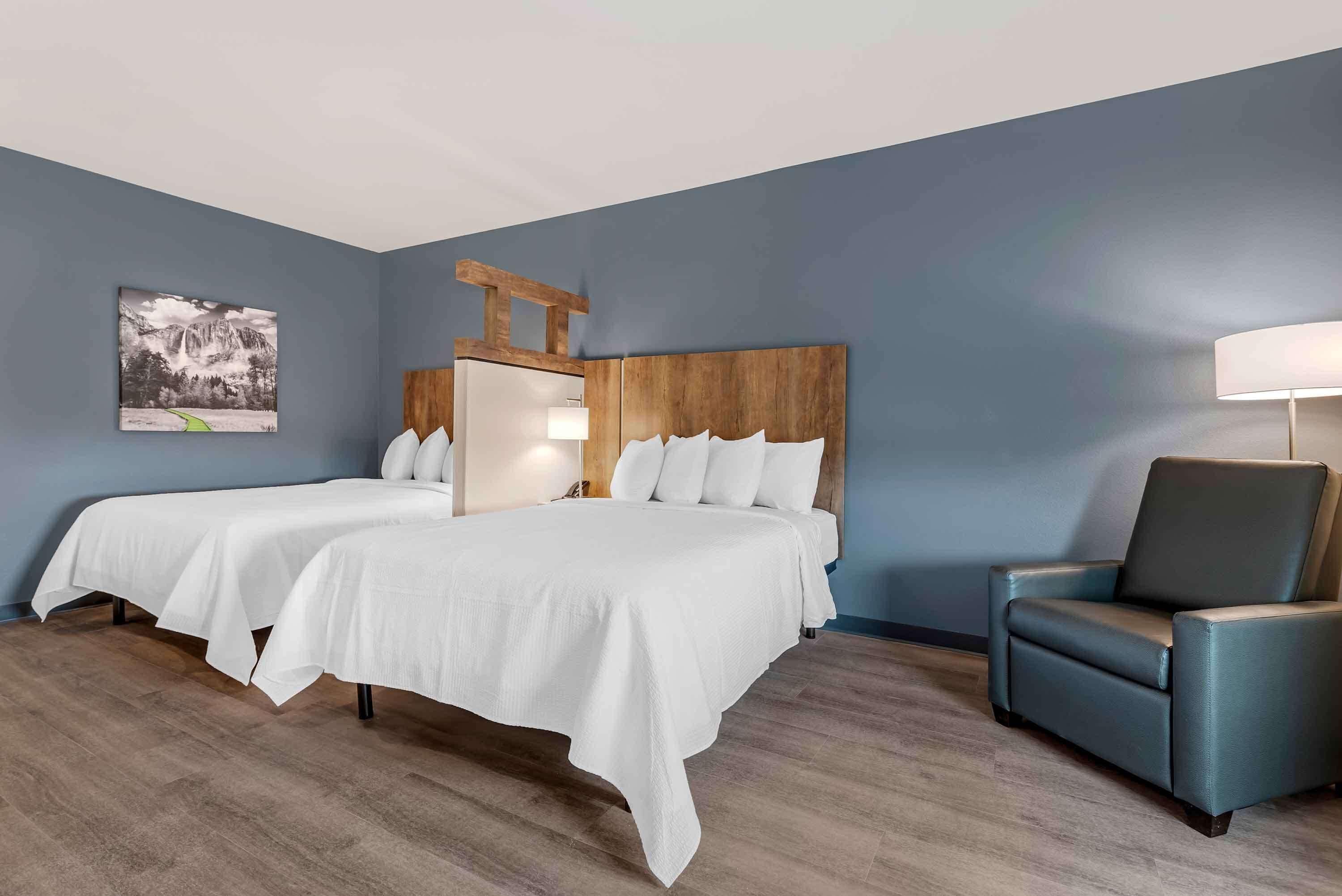 A hotel room with two beds, a wooden headboard, a blue armchair, and a painting on the wall.