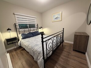 A bedroom with a bed, bedside lamps, a nightstand, a wooden cabinet, a window with blinds, and a framed picture on the wall. - Point Clark Cottage Getaway (Huron Kinloss)