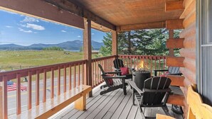 Une terrasse en bois avec des sièges d’extérieur, un foyer et une vue sur un paysage montagneux.