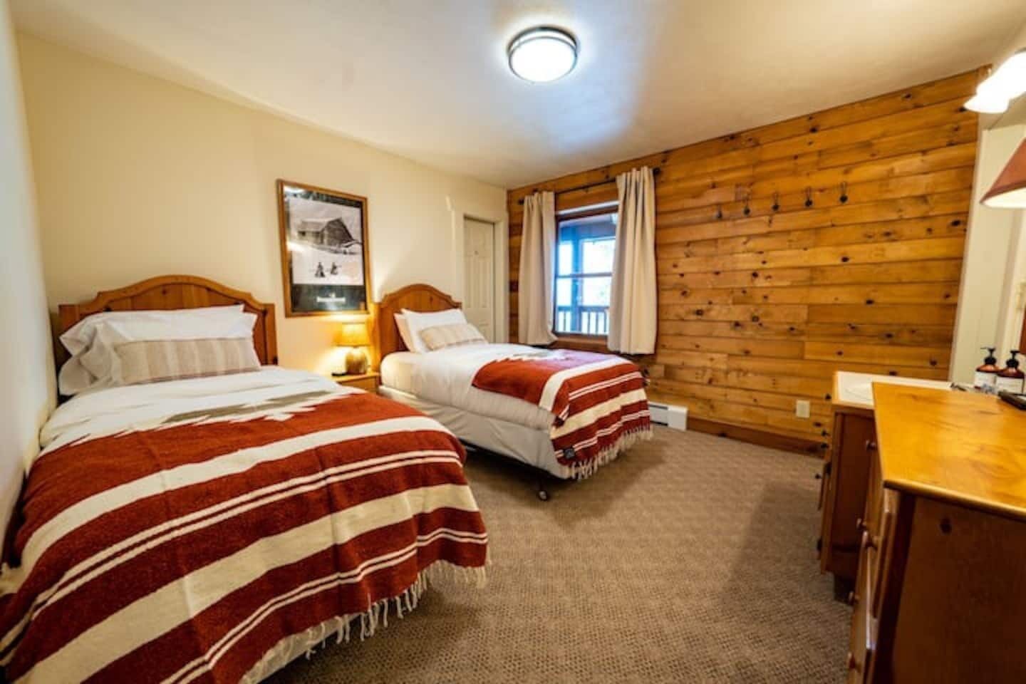 Une chambre avec deux lits, des murs en bois, un petit bureau et une fenêtre avec des rideaux.