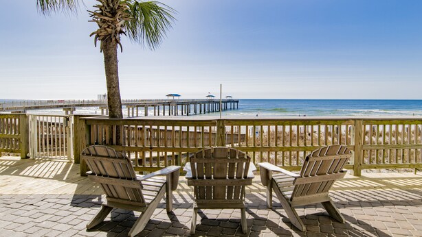 A wooden deck with two Adirondack chairs facing a beach, a palm tree, and a pier with umbrellas.