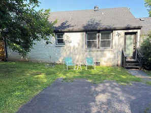 A house with a brick exterior, a front door, two windows, a small porch with a chair, and a grassy yard.