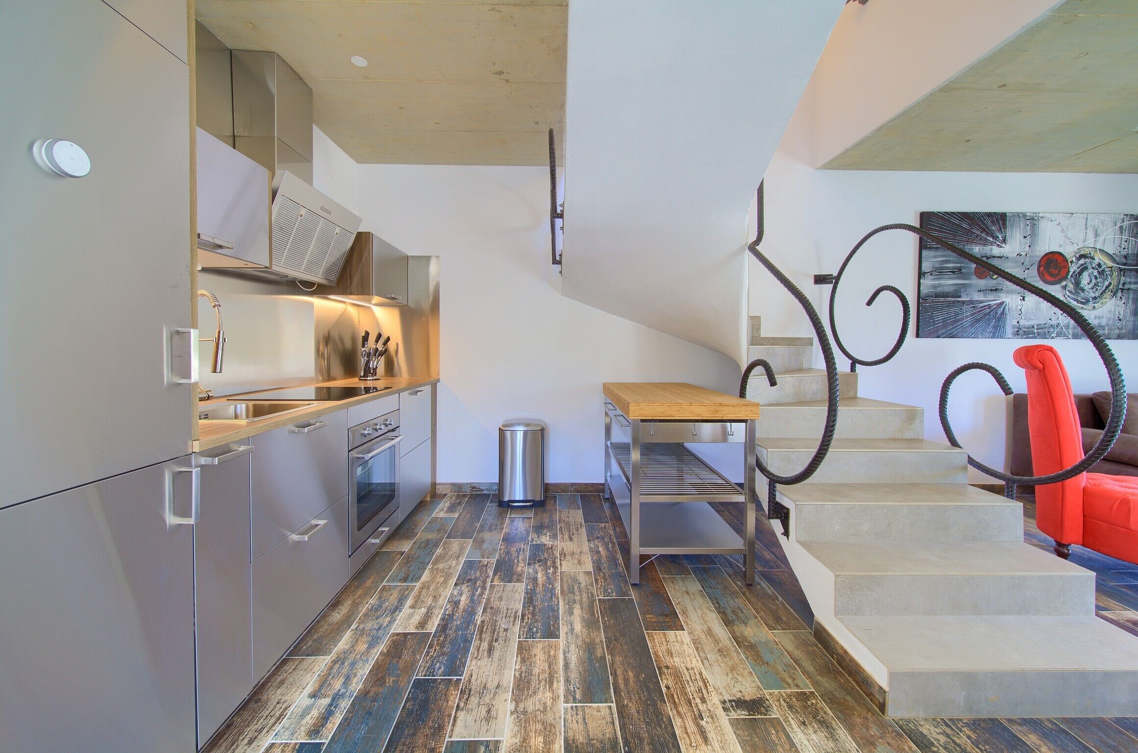 A modern kitchen with stainless steel appliances, a wooden table, and a staircase with a red accent.