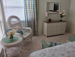 A bedroom with a wicker chair, a small table with a vase of flowers, a dresser with a mirror, and a bed with a quilt. - Runaway Bay 247 - Beach, pool, bay and gym (Bradenton Beach)