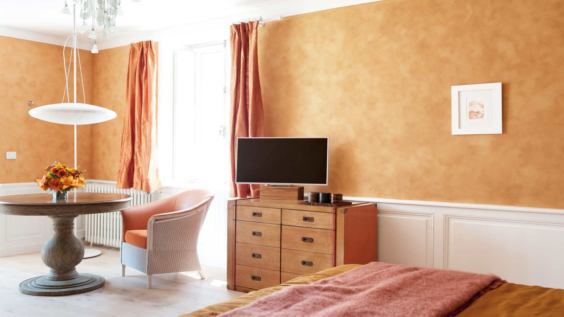 Camera d'albergo con un letto, una televisione, una cassettiera, una sedia, un tavolo con un vaso di fiori e un termosifone.