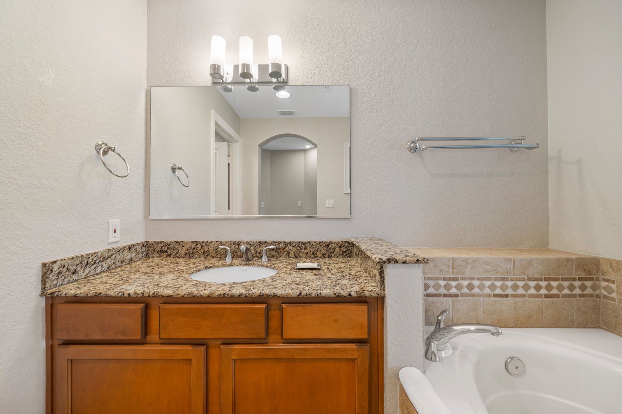 A bathroom with a granite countertop sink, wooden cabinets, a mirror with lights, a towel rack, and a bathtub.