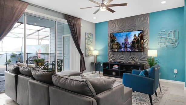 Un salon moderne avec un canapé gris, un fauteuil bleu, un téléviseur à écran plat fixé au mur et un ventilateur de plafond.