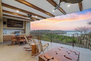 En rummelig balkon med udsigt over en solnedgang i en dal, møbler i træ og et moderne køkkenområde.