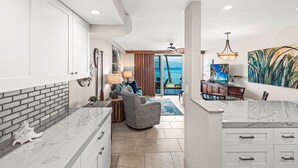 A spacious living area with a view of the ocean, a kitchen with white cabinets and a marble countertop, and a dining area with a table and chairs. - Luxury meets space in this Premium 1bd Oceanfront (Lahaina)